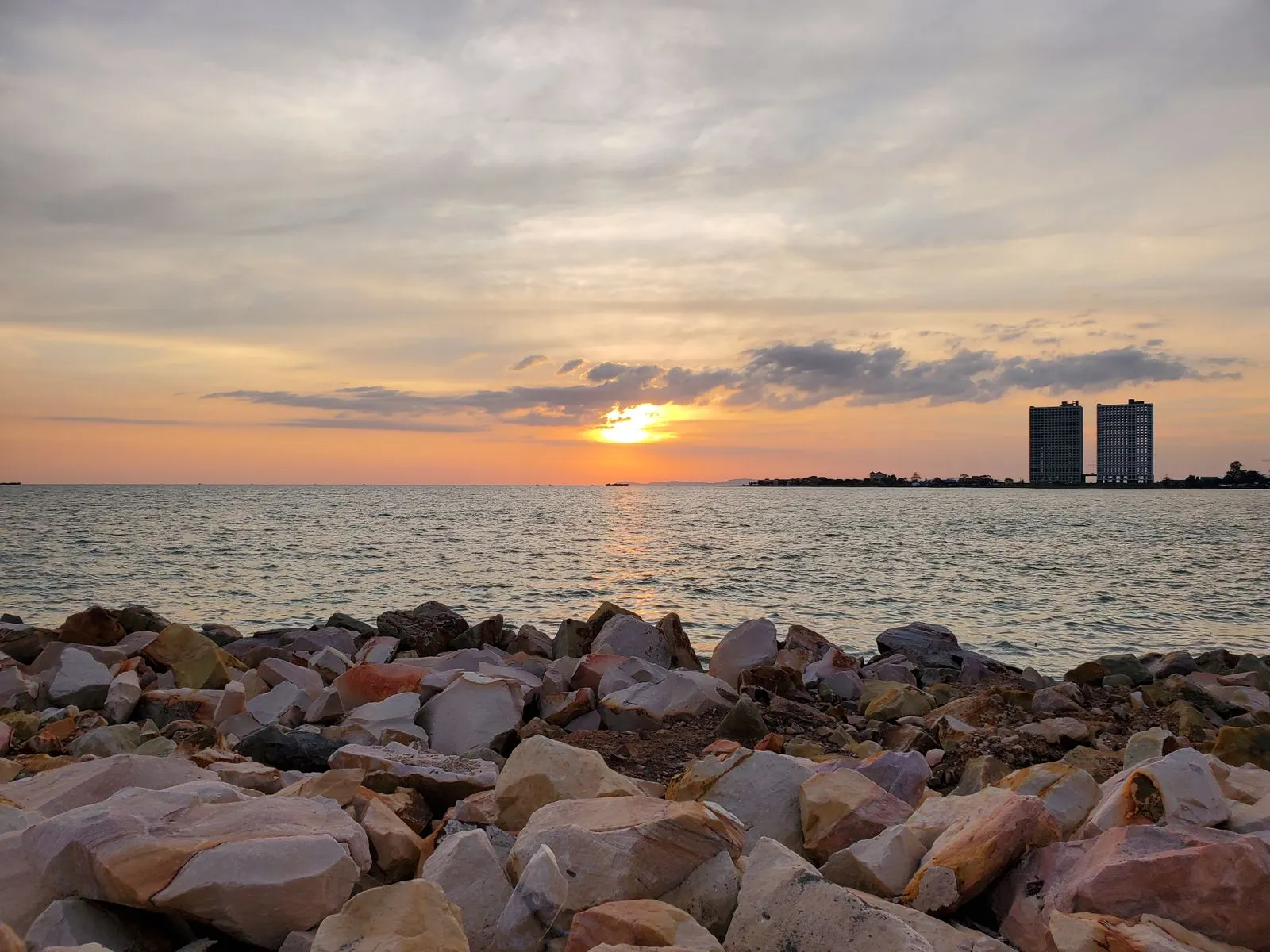 Beautiful sunset over the ocean with rocks in the foreground and buildings in the distance.